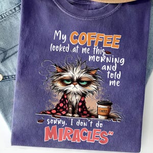 Puede incluir: Camiseta morada con un gato de dibujos animados y una taza de café. El texto de la camiseta dice "My coffee looked at me this morning and told me sorry, I don't do miracles".