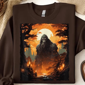 May include: A brown sweatshirt with a graphic print of a large, hairy Bigfoot standing in a forest setting. The background is a dark orange and brown color with a glowing orange moon.