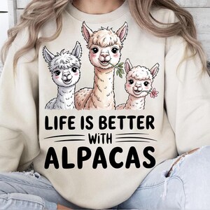 Op de afbeelding: Crèmekleurige sweatshirt met een afbeelding van drie lachende alpaca's, elk met een takje of bloem. De tekst "LIFE IS BETTER WITH ALPACAS" is eronder in het zwart gedrukt.