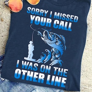 May include: A navy blue t-shirt featuring a graphic of a fisherman catching a fish. The text on the shirt reads "Sorry I missed your call I was on the other line".