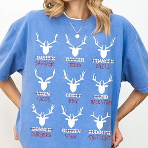 Puede incluir: Una camiseta azul claro con un gráfico blanco de cabezas de reno y nombres de alimentos tachados. La camiseta presenta las palabras "DASHER SAUSAGE", "DANCER JERKY", "PRANCER CHILI", "VIXEN TACOS", "COMET BBQ", "CUPID BACK STRAP", "DONNER BURGERS", "BLITZEN STEW" y "RUDOLPH NIGHT LIGHT".