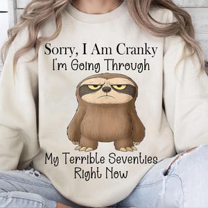 May include: Cream-colored sweatshirt with the text "Sorry, I Am Cranky I'm Going Through My Terrible Seventies Right Now." A cartoon sloth with a grumpy expression is centered below the text. The sloth is brown with a dark brown mask around its eyes.