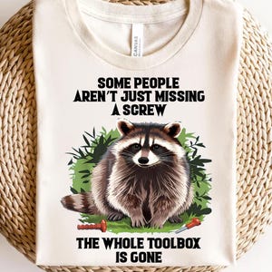 以下が含まれることがあります： ベージュのTシャツに、アライグマの黒いグラフィックと「Some people aren't just missing a screw. The whole toolbox is gone.」というテキストがプリントされています。