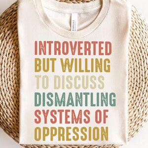 Könnte beinhalten: Ein weißes T-Shirt mit dem Text "Introvertiert, aber bereit, über die Demontage von Unterdrückungssystemen zu diskutieren" in einer Vintage-Schriftart. Der Text ist in Rot-, Gelb-, Grün- und Blautönen gehalten.