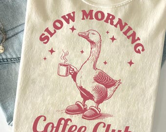 May include: Cream-colored t-shirt with a red graphic of a goose wearing slippers and holding a coffee cup. The text "SLOW MORNING Coffee Club" is arched above and below the goose illustration. The shirt is displayed on a light blue denim surface.