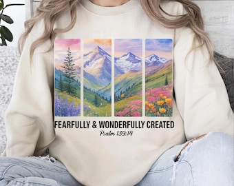 Puede incluir: Sudadera color crema con un diseño de paisaje con montañas, árboles y flores en paneles rectangulares. El texto "FEARFULLY & WONDERFULLY CREATED" y "Salmo 139:14" están impresos debajo. Una prenda superior cómoda e informal.