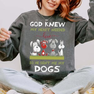 A woman wearing a gray sweatshirt with a graphic design of four cartoon dogs and text that reads "God Knew my heart needed love so he sent me my dogs".