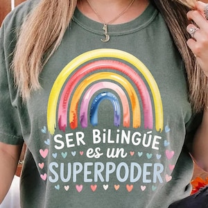 Könnte beinhalten: Grünes T-Shirt mit einem farbenfrohen Regenbogen-Motiv und dem spanischen Text „SER BILINGÜE es un SUPERPODER“ (Zweisprachig zu sein ist eine Superkraft). Das Regenbogen-Design zeigt gelbe, rote, rosa und blaue Bögen. Kleine Herzformen umgeben den Text.