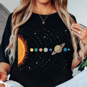 Puede incluir: Camiseta negra con un gráfico colorido del sistema solar. El diseño incluye un sol estilizado, planetas y estrellas. Los planetas se representan en varios colores, con Saturno y sus anillos.