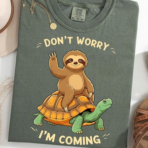 May include: A green t-shirt with a cartoon sloth riding a turtle. The text "Don't worry I'm coming" is printed on the shirt.