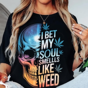 May include: Black t-shirt with a vibrant skull graphic and the words "I BET MY SOUL SMELLS LIKE WEED". The skull design uses blue, orange, and purple hues, with cannabis leaves integrated into the artwork.