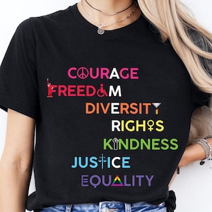 Könnte beinhalten: Schwarzes T-Shirt mit den Worten "COURAGE", "FREIHEIT", "VIELFALT", "RECHTE", "GÜTE", "GERECHTIGKEIT" und "GLEICHHEIT" in Regenbogenfarben bedruckt.