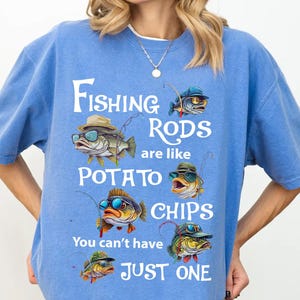 May include: A light blue t-shirt with the text "Fishing Rods are like Potato Chips, You can't have JUST ONE." The shirt features cartoon fish wearing hats and sunglasses, with fishing rods. The shirt is a comfortable, casual style.