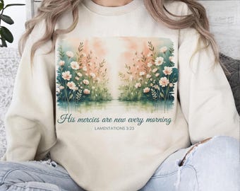 May include: Cream-colored sweatshirt featuring a watercolor floral design with the text "His mercies are new every morning" from Lamentations 3:23. The artwork depicts a serene landscape with flowers and a reflective body of water, in shades of pink, green, and blue.