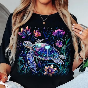 May include: Black t-shirt featuring a vibrant, artistic illustration of a sea turtle surrounded by colorful flowers and aquatic plants. The turtle's shell is a mosaic of blues, purples, and greens. The flowers include pink, purple, and orange hues.