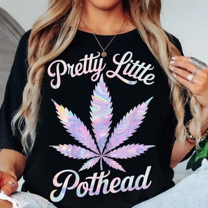 May include: Black t-shirt with the text "Pretty Little Pothead" in white cursive. A large, colourful cannabis leaf graphic is centered below the text, with shades of purple, blue, and pink.