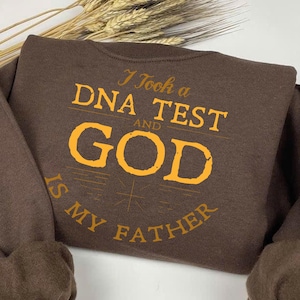 Puede incluir: Camiseta de manga larga marrón con un gráfico amarillo que dice "Hice una prueba de ADN y Dios es mi Padre".