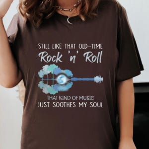 Puede incluir: Una camiseta marrón con un gráfico de una guitarra con una luna en el agujero de sonido. El texto en la camiseta dice "Still like that old-time Rock 'n' Roll That kind of music just soothes my soul."