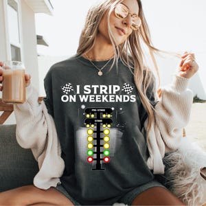 Puede incluir: Sudadera gris oscuro con el texto "I Strip on Weekends" y un gráfico de árbol de Navidad de carreras de resistencia. El gráfico presenta luces amarillas, verdes y rojas. Dos banderas a cuadros están a cada lado del texto.