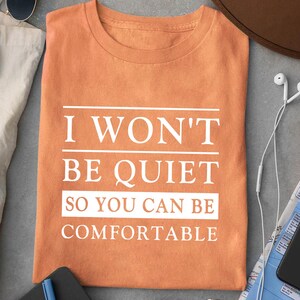 May include: Orange t-shirt with the text "I won't be quiet so you can be comfortable" printed in white.
