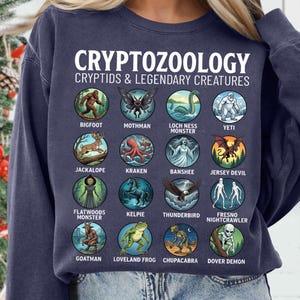 May include: A navy blue crewneck sweatshirt featuring a white graphic print titled CRYPTOZOOLOGY CRYPTIDS & LEGENDARY CREATURES. The design displays sixteen circular illustrations of various mythical creatures with their names printed below each icon.