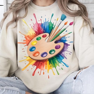 Pode incluir: Uma camisola creme com um design de paleta de pintura colorida. O design inclui uma paleta de madeira com várias cores de tinta, pincéis e um vibrante efeito de respingos de arco-íris. É uma peça de roupa casual.
