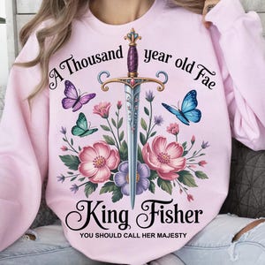 Könnte beinhalten: Ein hellrosa Sweatshirt mit einem Blumen- und Schmetterlingsdesign und einem Schwert. Der Text lautet "A Thousand year old Fae" und "King Fisher, You should call her Majesty".