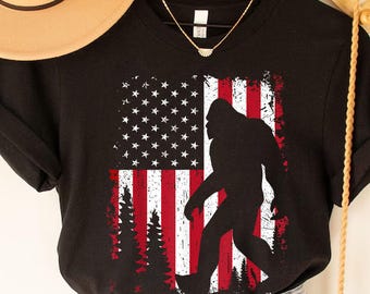 Puede incluir: Camiseta negra con un diseño desgastado de la bandera estadounidense y una silueta de Bigfoot. El diseño de la bandera incluye estrellas blancas y rayas rojas. La camiseta es una prenda informal. El diseño está impreso en la parte delantera.