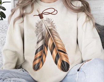 May include: Cream-colored sweatshirt featuring a detailed illustration of two feathers. The feathers are in shades of brown, black, and white, with a brown leather-like tie at the top. The design has a Native American or bohemian aesthetic.