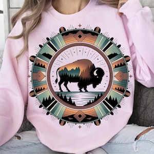 May include: A light pink sweatshirt featuring a circular design with a bison silhouette. The bison is set against a landscape of mountains and water, framed by geometric patterns in shades of green, brown, and orange.