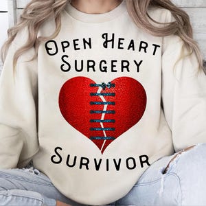 May include: Cream-colored sweatshirt with the text "Open Heart Surgery Survivor" in black. A large red heart with blue stitching is centered on the front. The heart design has a textured appearance.