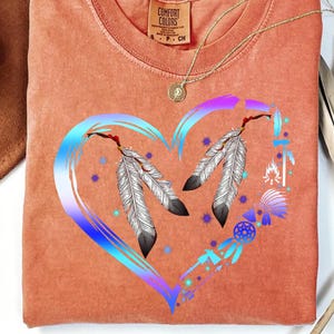 May include: A light orange t-shirt with a blue and purple heart design. The heart features two white feathers and a dreamcatcher with arrows. The text "COMFORT COLORS" is visible on the shirt.