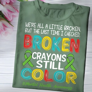 May include: Green t-shirt with the text "We're all a little broken. But the last time I checked, broken crayons still color." and the hashtag "#Mental Health Awareness".