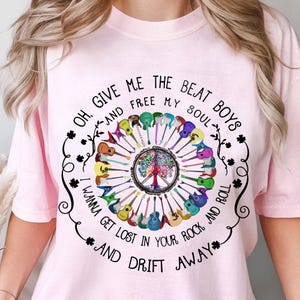 May include: Pink t-shirt with a black graphic design featuring a peace sign surrounded by colorful guitars and the text "Oh, give me the beat boys and free my soul I wanna get lost in your rock and roll and drift away".