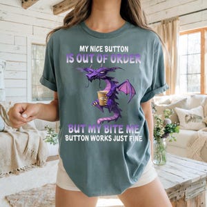 May include: A slate blue t-shirt featuring a graphic of a purple dragon holding a coffee cup. The text reads: MY NICE BUTTON IS OUT OF ORDER BUT MY BITE ME BUTTON WORKS JUST FINE. The design is centered on the chest with white and purple lettering.