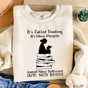 以下が含まれることがあります： クリーム色のスウェットシャツに、本を読んでいる女の子が本の上に座っている黒いグラフィックが描かれています。テキストには「It's Called Reading It's How People Install New Software Into Their Brains」と書いてあります。