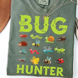 May include: A green t-shirt with the words "BUG HUNTER" in large, lime green letters. The shirt features cartoon illustrations of various insects, including an ant, a caterpillar, a snail, a spider, a ladybug, and a bee.