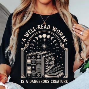 May include: Black t-shirt with a graphic design featuring books, celestial elements, and the text "A WELL-READ WOMAN IS A DANGEROUS CREATURE". The design includes a crescent moon phase and stars.