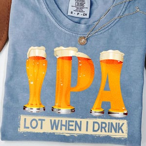 May include: A pale blue t-shirt featuring a beer-themed graphic. The word "IPA" is formed by three beer glasses, with the text "LOT WHEN I DRINK" below. A silver necklace with a pendant is also visible.