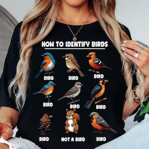 Może przedstawiać: Czarny t-shirt z kolorową ilustracją różnych ptaków i napisem "HOW TO IDENTIFY BIRDS". Każdy ptak jest oznaczony jako "BIRD", z wyjątkiem wiewiórki oznaczonej jako "NOT A BIRD".
