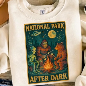 May include: Cream-colored sweatshirt with a graphic featuring an alien, Bigfoot, and a bear roasting marshmallows around a campfire under a full moon. The graphic is framed with the words "NATIONAL PARK AFTER DARK" in gold lettering.