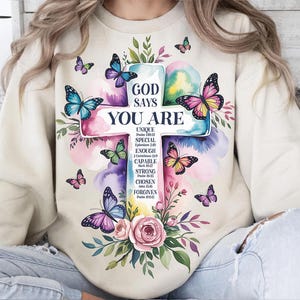May include: A cream-colored sweatshirt featuring a cross design with the words "GOD SAYS YOU ARE." The cross is decorated with colorful butterflies and floral elements. The sweatshirt has long sleeves and a crew neck.