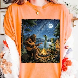 May include: Orange t-shirt featuring a whimsical illustration of Bigfoot playing a guitar by a campfire with an alien and a lake monster. The scene is set at night under a full moon and starry sky.