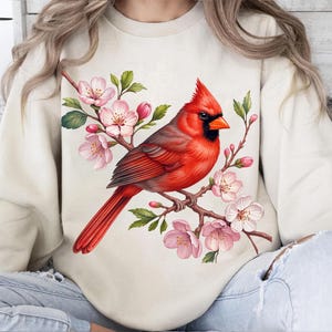 May include: A cream-colored sweatshirt featuring a vibrant illustration of a red cardinal perched on a flowering branch. The bird has black accents and an orange beak. The blossoms are pink and white, with green leaves.