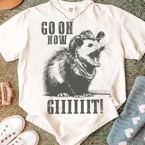 Può includere: T-shirt color crema con grafica di un opossum con cappello da cowboy e bandana, con la scritta "GO ON NOW GIIIIIIIT!". L'opossum è raffigurato con la bocca aperta. La maglietta ha una vestibilità ampia.