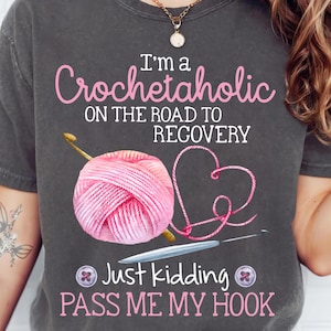 Può includere: Una maglietta grigia con testo rosa che recita "Sono una crochet-dipendente sulla strada della guarigione Scherzo, passa il mio uncinetto". La maglietta presenta anche un gomitolo di lana rosa e un uncinetto.