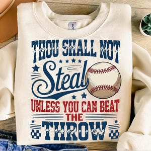 May include: Cream-colored sweatshirt with the text "THOU SHALL NOT STEAL UNLESS YOU CAN BEAT THE THROW" in navy blue and red. A baseball graphic is featured on the front. The sweatshirt has a crew neck and long sleeves.