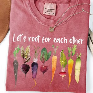 Op de afbeelding: Een roze T-shirt met de tekst "Let's root for each other" in wit gedrukt. Het shirt heeft een rij kleurrijke groenten, waaronder wortels, radijsjes en bieten.