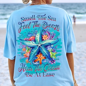 Può includere: Una maglietta blu chiaro con un design a stella marina colorato e il testo "Smell The Sea Feel The Breeze Hear The Ocean Be At Ease".