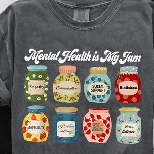 May include: Dark grey t-shirt with the slogan "Mental Health is My Jam." The design features eight illustrated jam jars, each labelled with a mental health concept, including "Empathy" and "Mindfulness."